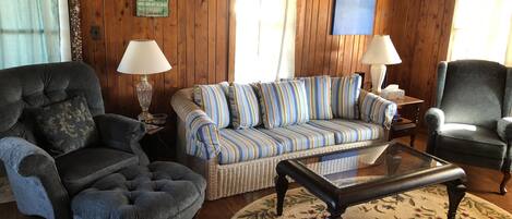 A cozy living room with wooden walls, a striped sofa, two armchairs, a glass coffee table, and a floral rug.