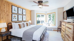 Un dormitorio con cama, ventilador de techo, impresiones enmarcadas de motivos botánicos, televisor y ventana con vistas a la vegetación.