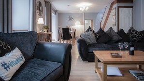 A modern living room with a sofa, a coffee table, and a dining area in the background. - Rose Coach House - 3 Bedroom Cottage - Pendine (Carmarthen)