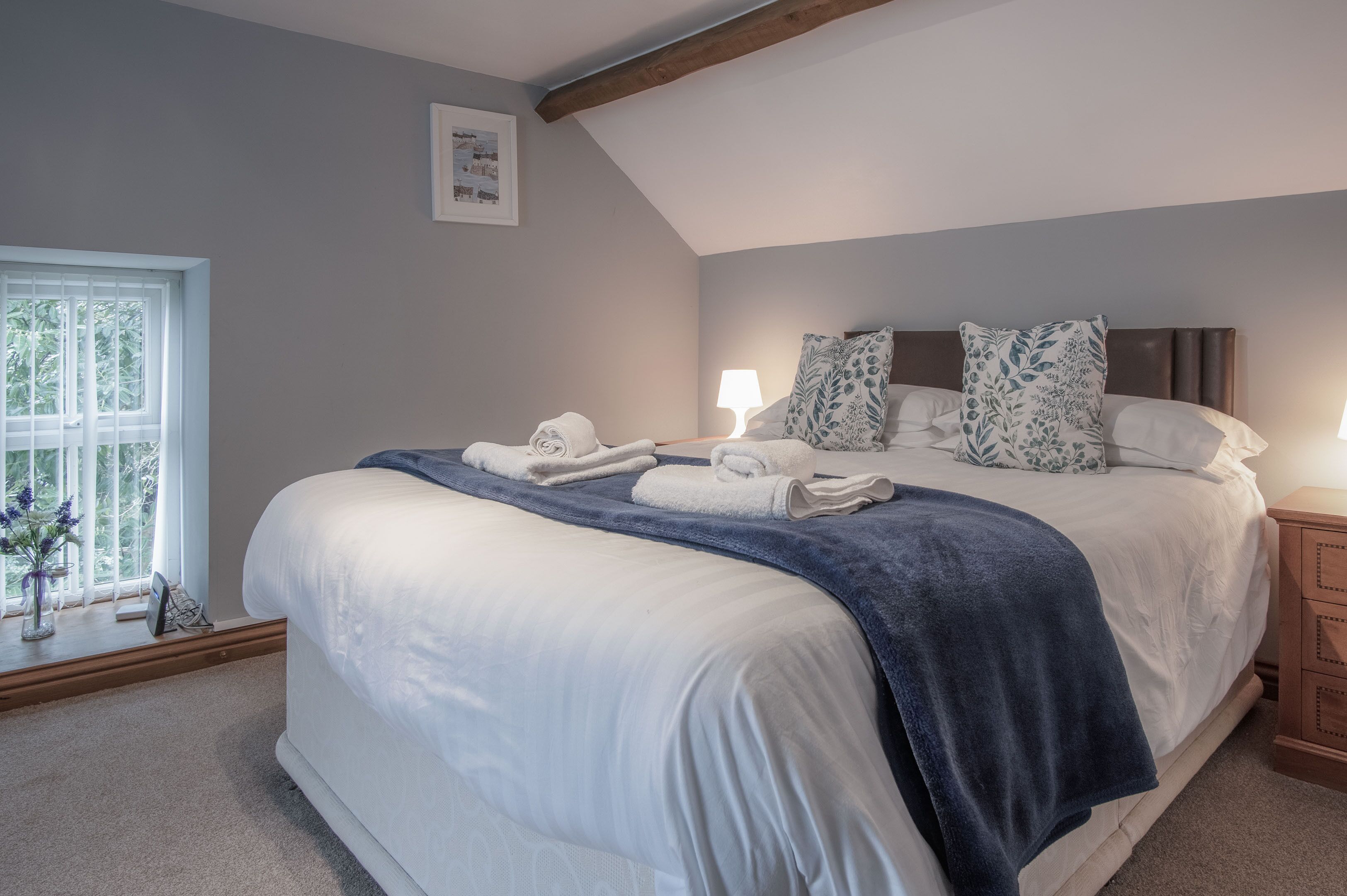 Een net opgemaakt bed met witte lakens en een blauwe deken, een bedlampje en een raam met uitzicht op bomen.