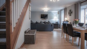 A modern living room with a grey sofa, wooden staircase, and a dining area with a table and chairs. - Rose Coach House - 3 Bedroom Cottage - Pendine (Carmarthen)