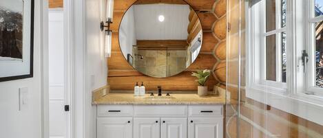 A bathroom with a wooden wall, a round mirror, a sink with two bottles, and a potted plant.