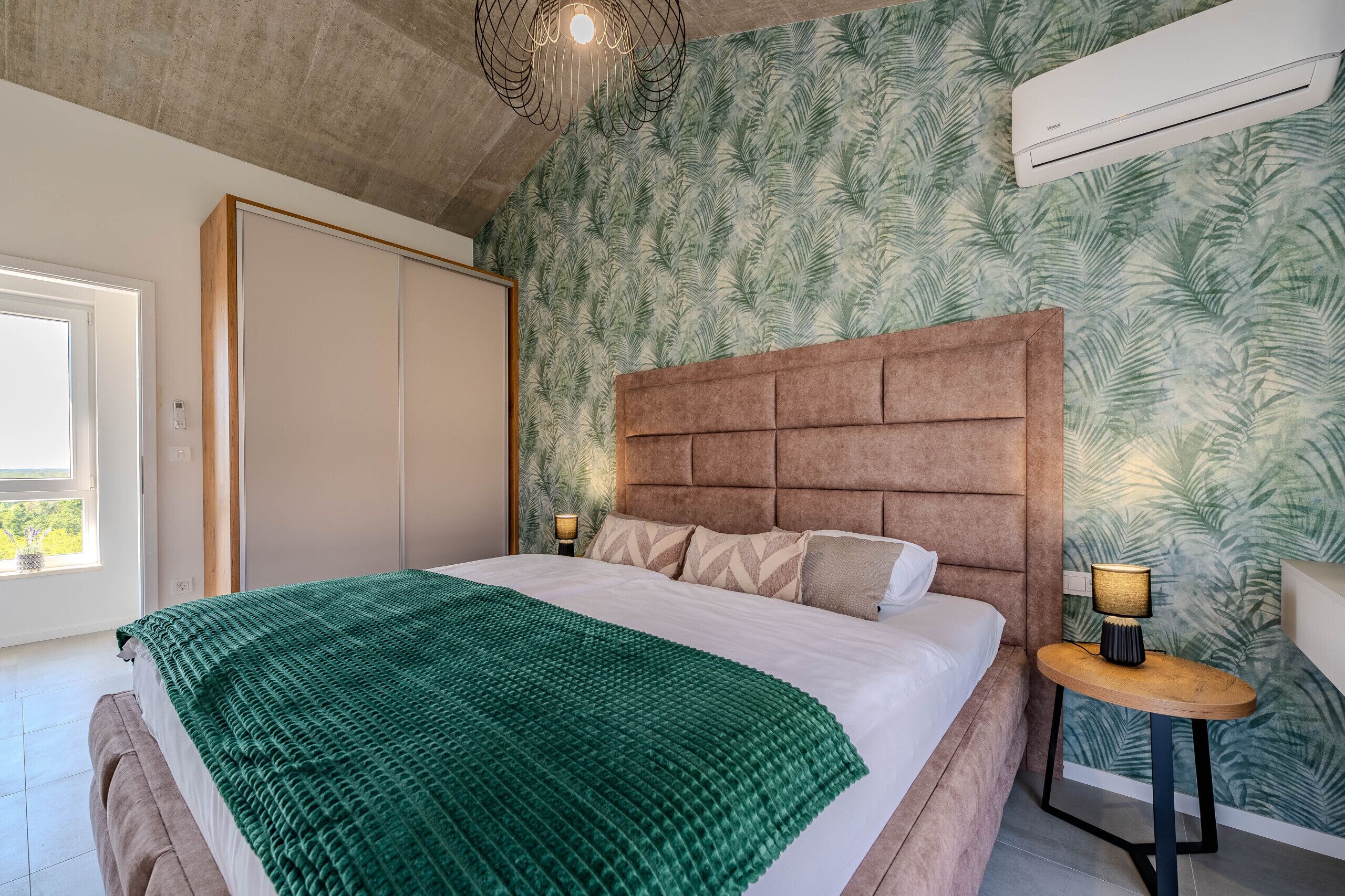 A bedroom with a large bed, a headboard, a nightstand, a lamp, and a window.