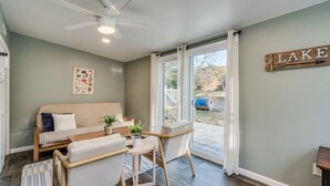 Un salon confortable avec un canapé, des fauteuils, une petite table et un ventilateur de plafond. Une fenêtre avec des rideaux donne sur une terrasse équipée d’un barbecue.