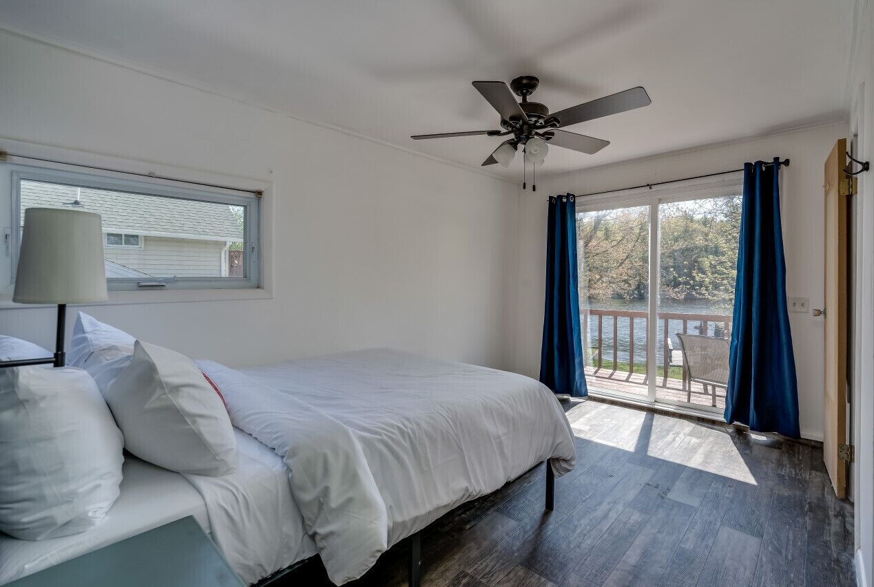 Un dormitorio con cama, ventilador de techo, ventana y vista a un balcón con barandilla.