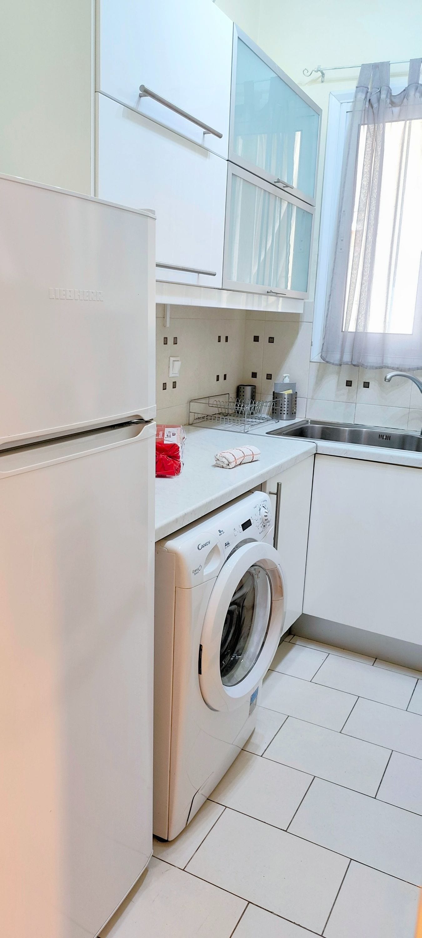 A modern kitchen with white cabinets, a built-in washing machine, a stainless steel sink, and a window with sheer curtains.