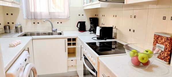 A compact kitchen with a sink, stove, and a coffee maker. There is a bowl of fruit on the counter and a cereal container on the wall.