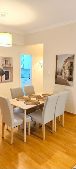Uma sala de jantar com uma mesa posta para quatro pessoas, cadeiras em tons claros, um lustre e uma luminária de parede, além de um quadro com uma cena de rua.