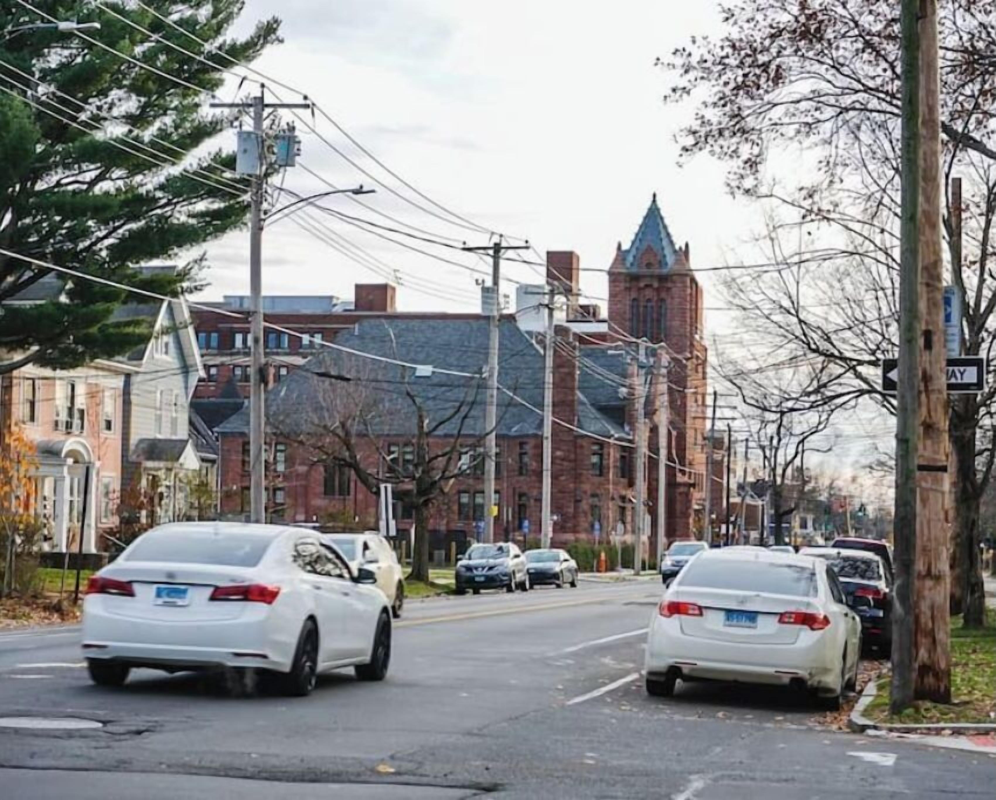 A street view with cars parked on the side, a building with a tower, and power lines overhead.