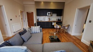 A modern living room with a grey sectional sofa, a glass coffee table, and a kitchen area with dark cabinets and a microwave.