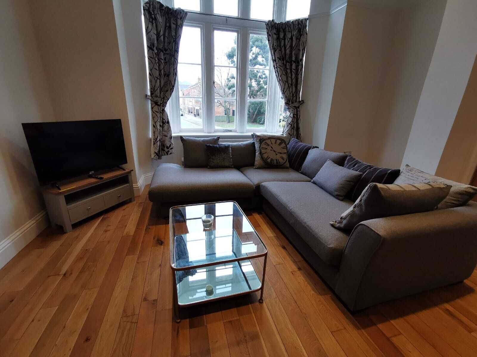 A living room with a sectional sofa, a glass coffee table, a flat-screen TV, and a window with curtains.