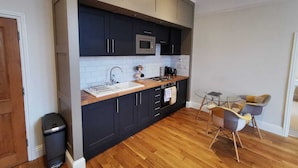 A modern kitchen with dark cabinets, a wooden countertop, a sink, and a microwave. There is a small table with chairs and a trash bin.