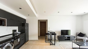 A modern kitchen with dark cabinets, a built-in oven, and a washing machine. There is a dining area with a TV, a sofa, and a coffee table. - Sunset 302 Apt de Luxe Avec Salle de Fitness (Tunis)