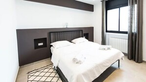 A modern hotel room with a bed, a bedside table, a window with curtains, and a wall-mounted light fixture. - Sunset 302 Apt de Luxe Avec Salle de Fitness (Tunis)