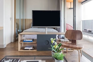 A modern bedroom with a bed, a TV, a desk with books, and a plant.