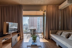 Una sala de estar moderna con un televisor de pantalla plana, un sofá, una mesa de centro con una planta y un balcón con mesa y sillas.