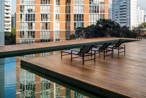A rooftop pool with wooden decking and lounge chairs, surrounded by a high-rise building with orange and white brick facade.