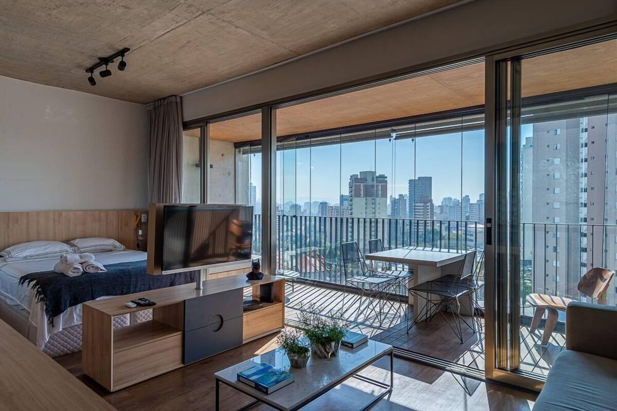 Una habitación de hotel moderna con un balcón grande, una cama, un televisor, un escritorio y un rincón de comedor.
