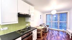 A compact kitchen with a stove, oven, and white cabinets. A dining area with a table and chairs is visible, and a sofa is placed near a window with blinds. - Lovely 1-bed Apartment in London by the Harbour (London)