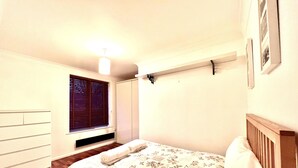 A bedroom with a bed, a window with blinds, a bedside table, and a wall-mounted shelf. - Lovely 1-bed Apartment in London by the Harbour (London)