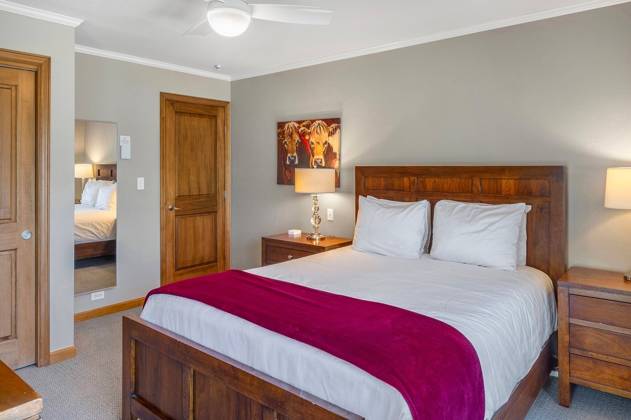 Un dormitorio con una cama de madera, una manta roja, una mesita de noche con lámpara, una puerta de madera y un cuadro colgado en la pared.