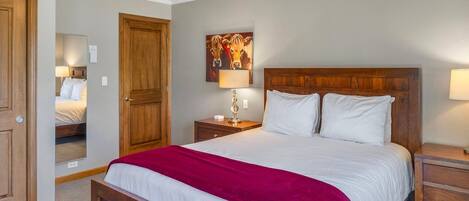 Un dormitorio con una cama de madera, una manta roja, una mesita de noche con lámpara, una puerta de madera y un cuadro colgado en la pared.