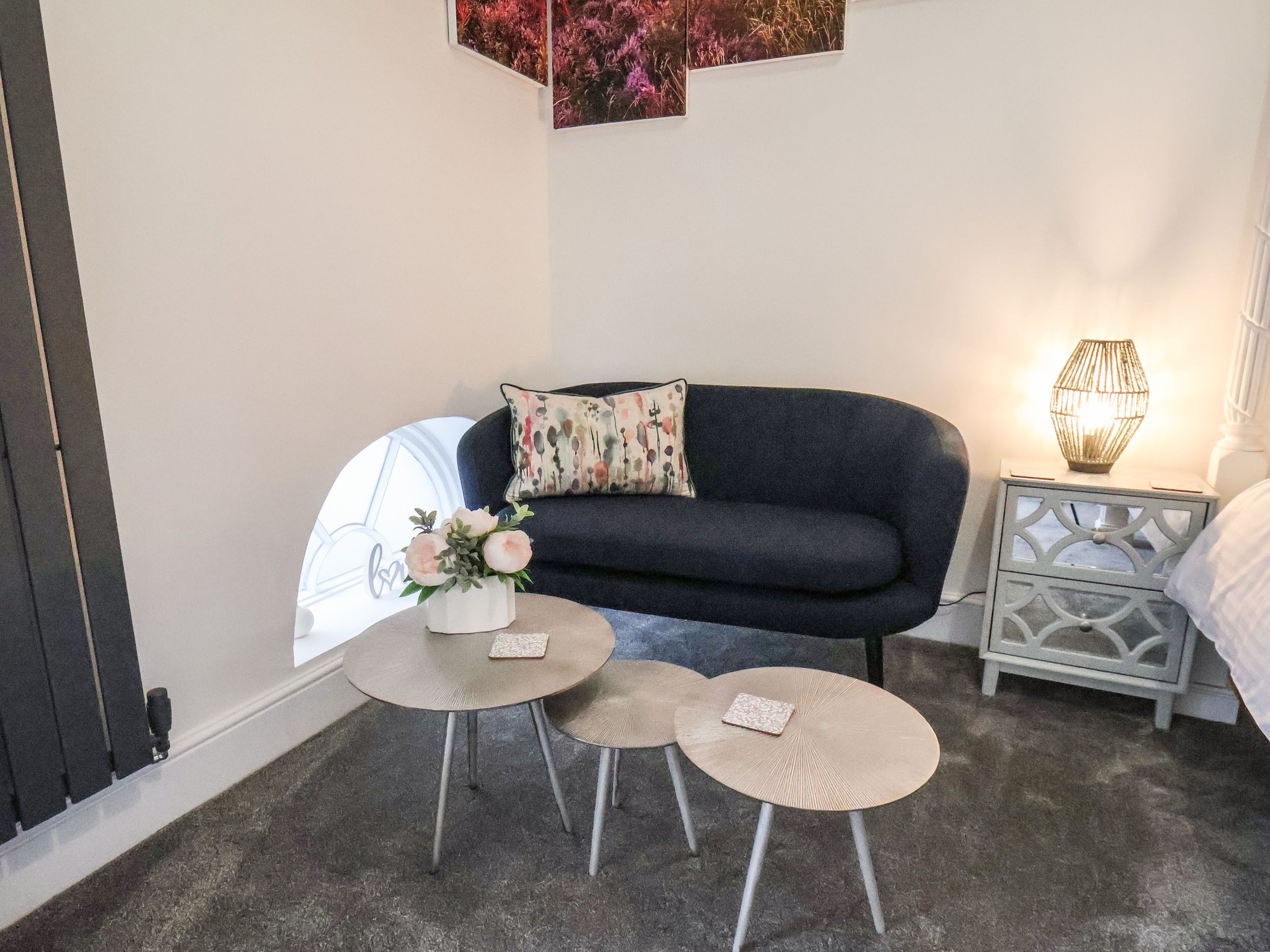 A cozy room with a dark sofa, two small round tables, a decorative pillow, a wall-mounted artwork, and a bedside lamp.