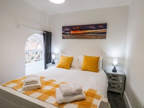 A neatly made bed with white and yellow bedding, two bedside lamps, and a painting of a sunset over a pier on the wall. - Riverside Chapel (Middlesbrough)
