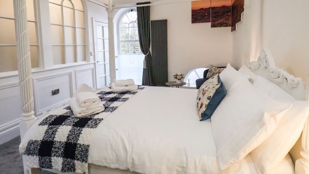 Una cama con dosel con una colcha a cuadros, una mesita redonda con un florero, y vista a un atardecer a través de una ventana.