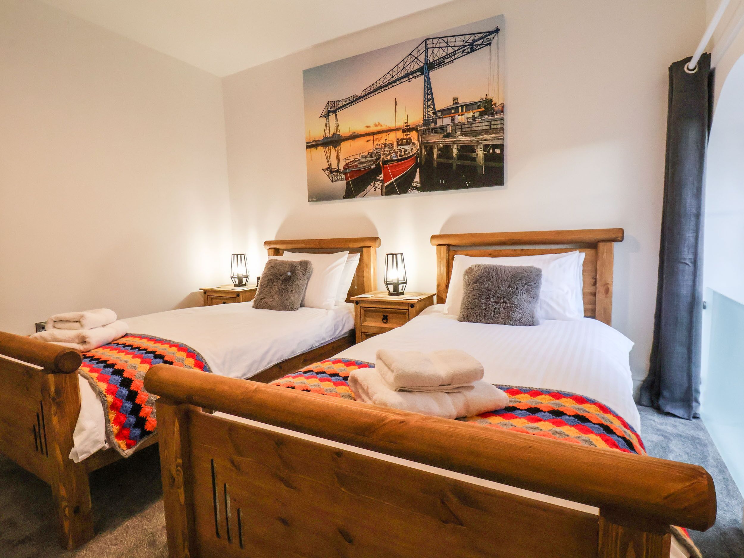 A hotel room with two wooden beds, each with a headboard, white linens, and a colorful blanket. There is a painting of a bridge on the wall above the beds.