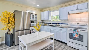 A modern kitchen with white cabinets, stainless steel appliances, and a white table with a vase of yellow flowers. - Galveston Retreat w/ Backyard & Patio! (Galveston)
