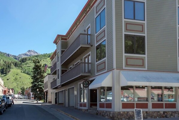 Un edificio de varios pisos con balcones, un escaparate comercial y montañas al fondo.