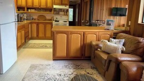 A compact kitchen with wooden cabinets and a central island, a living area with a leather sofa and a rug, and a television mounted on the wall. - *Ten Trees* Cabin, Secluded, Near Redwood Parks (Myers Flat)