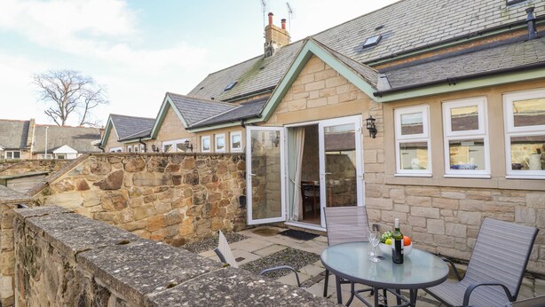 A stone-walled patio with a round table and chairs, a stone wall, and a building with a tiled roof and windows. - Willow Cottage (Alnwick)