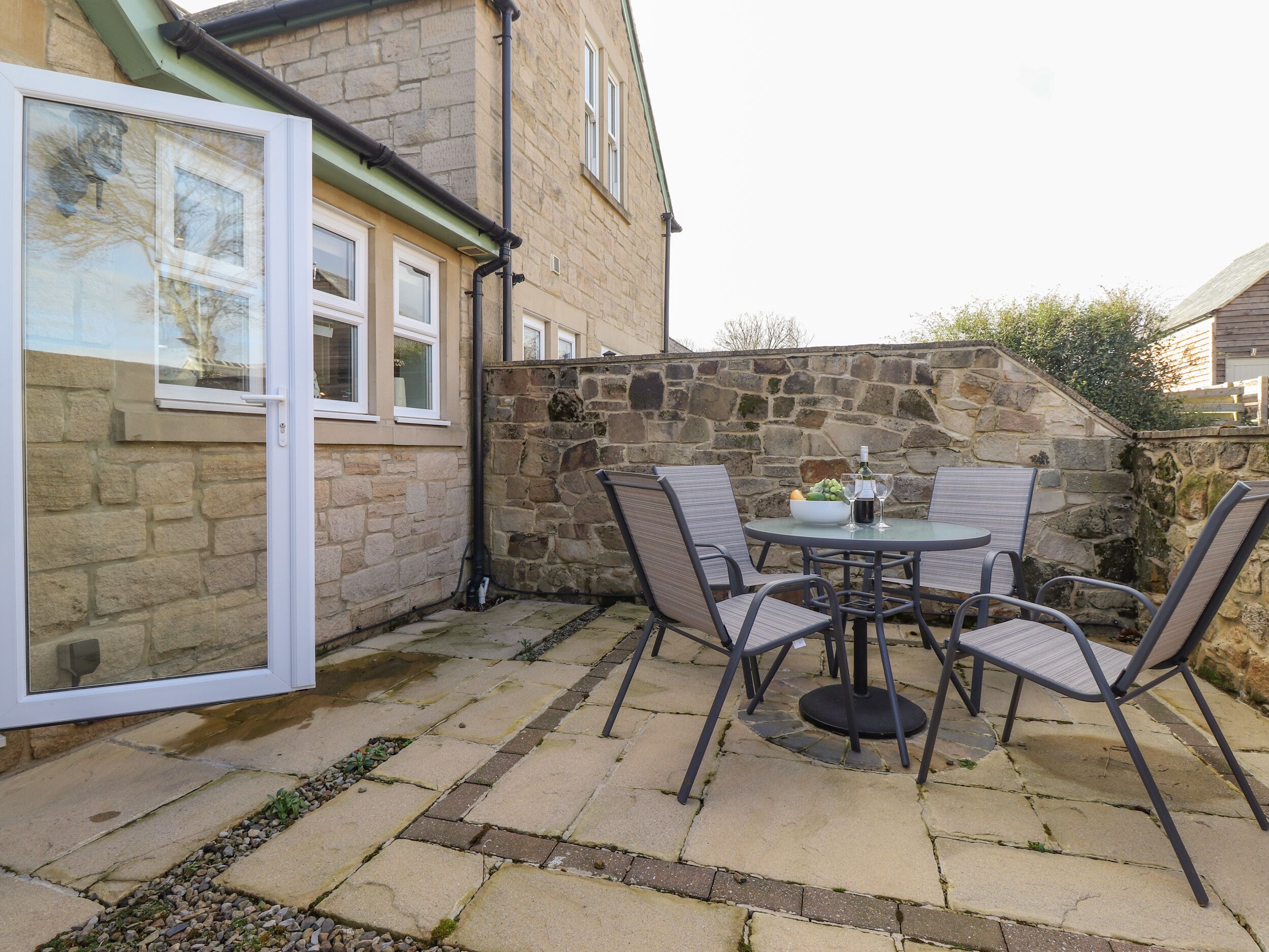 An outdoor patio with a stone wall, a table with chairs, and a glass door leading to a room.
