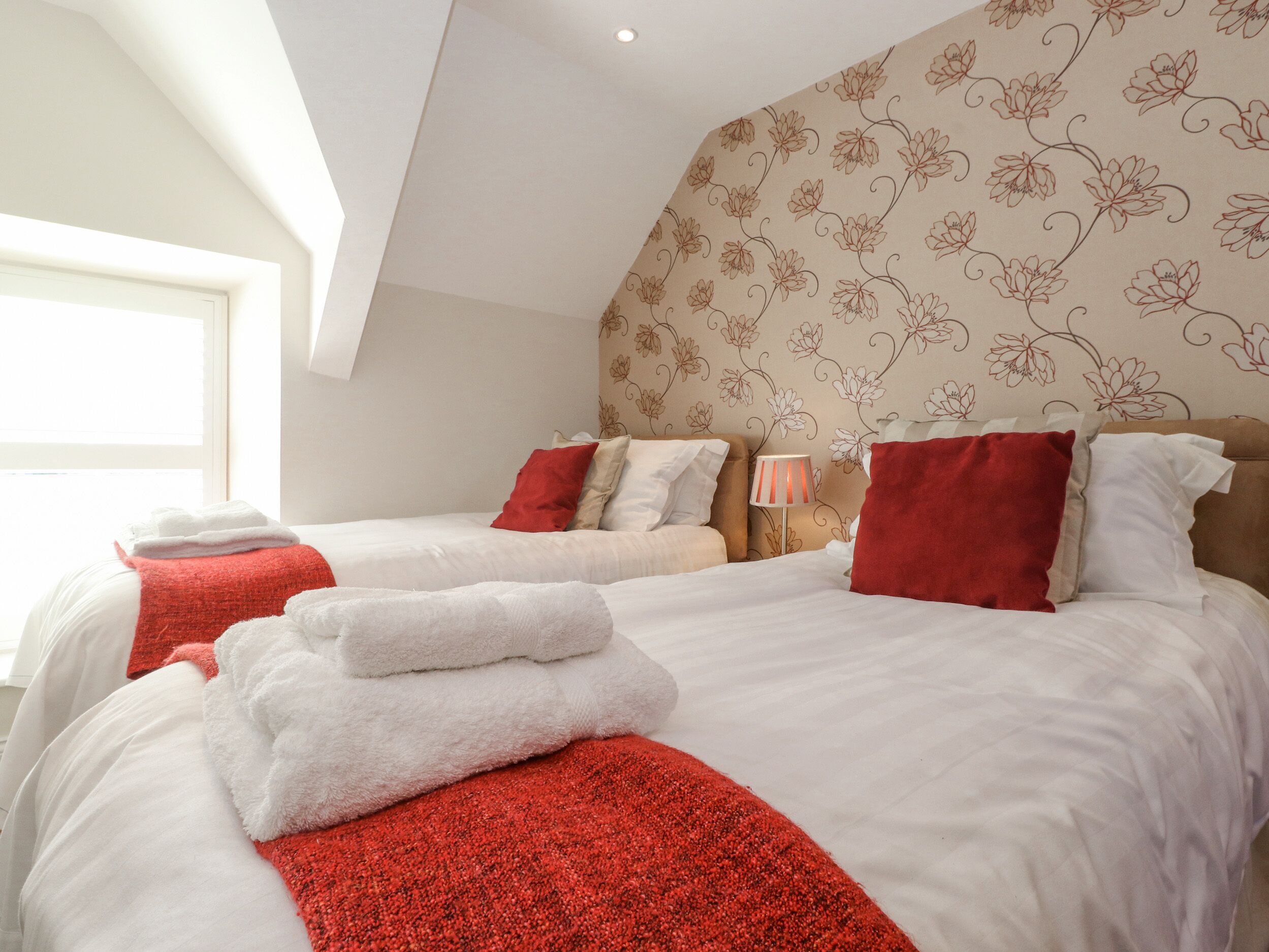 A hotel room with two beds, red and white bedding, a window, and floral wallpaper.