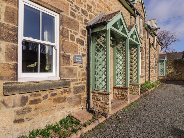 A stone cottage with a window, a door with a lattice design, and a gravel path. - Willow Cottage (Alnwick)