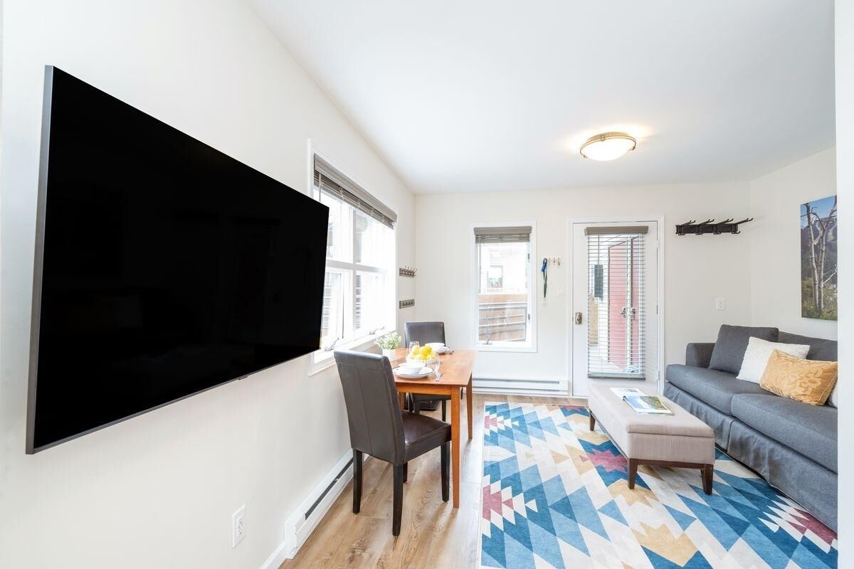 Uma sala de estar moderna com uma TV de tela plana na parede, uma mesa de jantar com cadeiras, um sofá cinza e um tapete colorido.