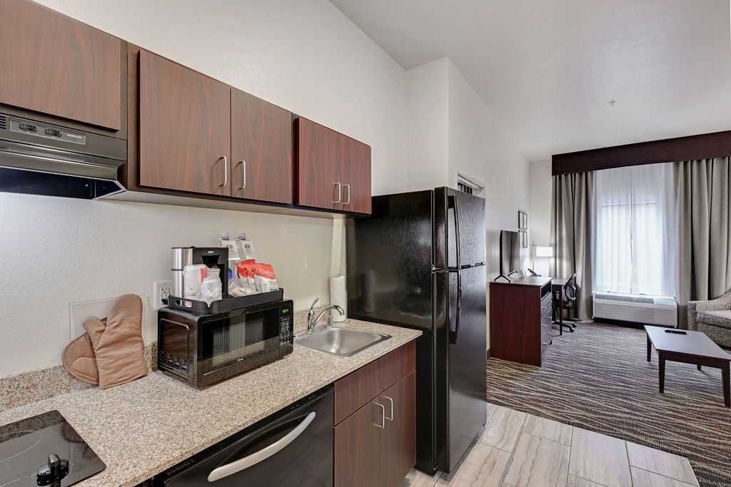 A modern kitchen with a built-in microwave, oven, and refrigerator. There is a sink and a coffee maker on the counter. The room has a large window with curtains and a seating area with a sofa and a small table.