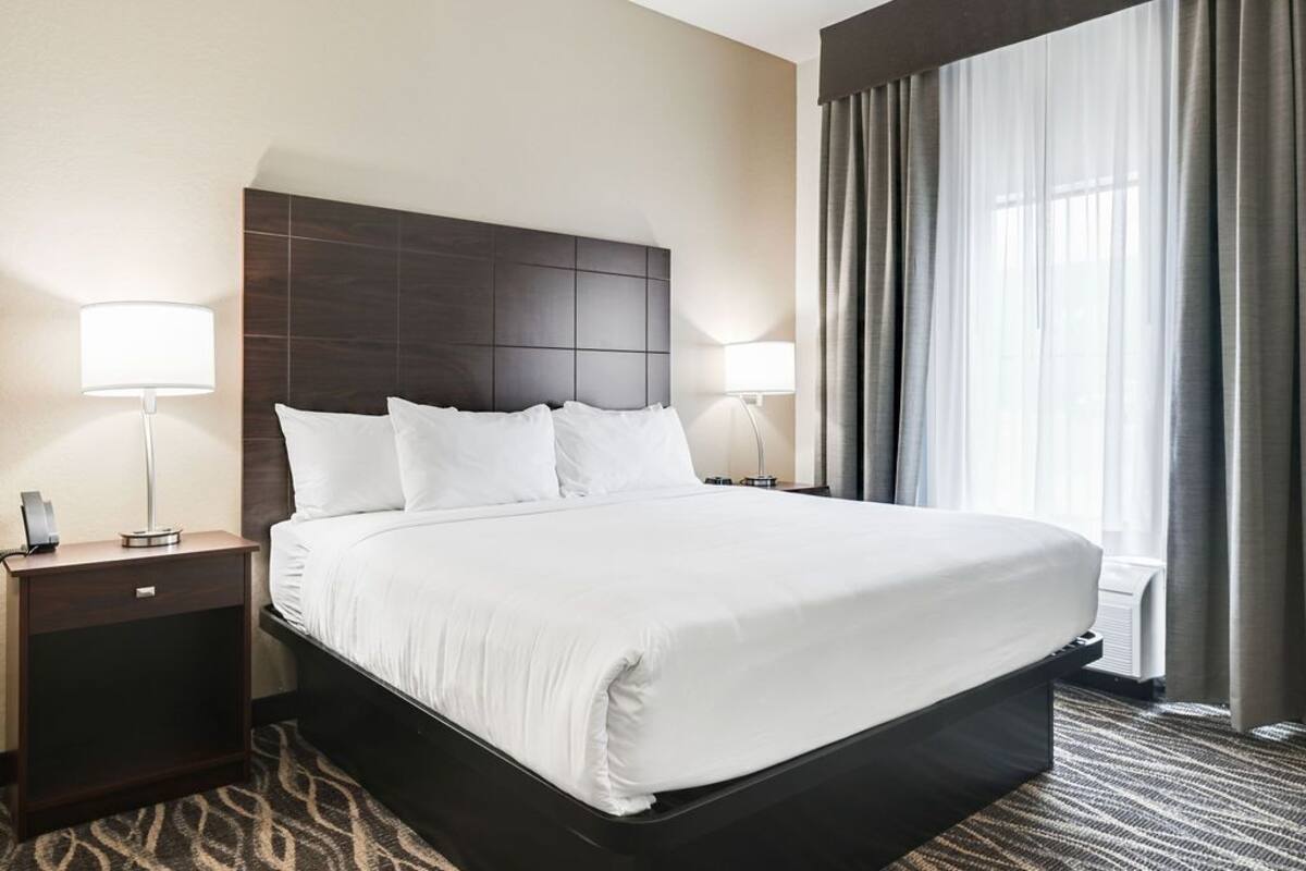 Une chambre d’hôtel avec un grand lit, deux tables de chevet, une tête de lit et une fenêtre avec des rideaux.
