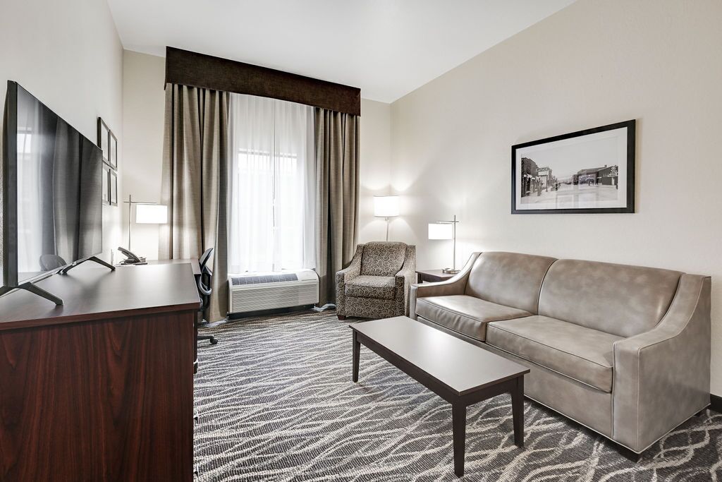 Habitación de hotel con escritorio, sofá, silla, mesita de centro, ventana grande con cortinas, lámpara de pie y un cuadro en la pared.