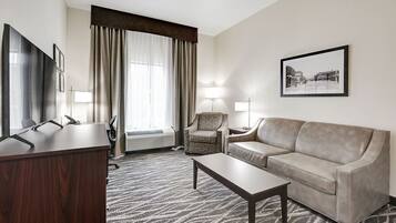 Habitación de hotel con escritorio, sofá, silla, mesita de centro, ventana grande con cortinas, lámpara de pie y un cuadro en la pared.