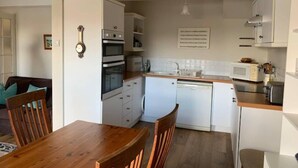 A compact kitchen with white cabinets, a microwave, a dishwasher, and a wooden dining table with chairs. - Newly Refurbished 7-bed Cottage in Polruan, Fowey (Fowey)