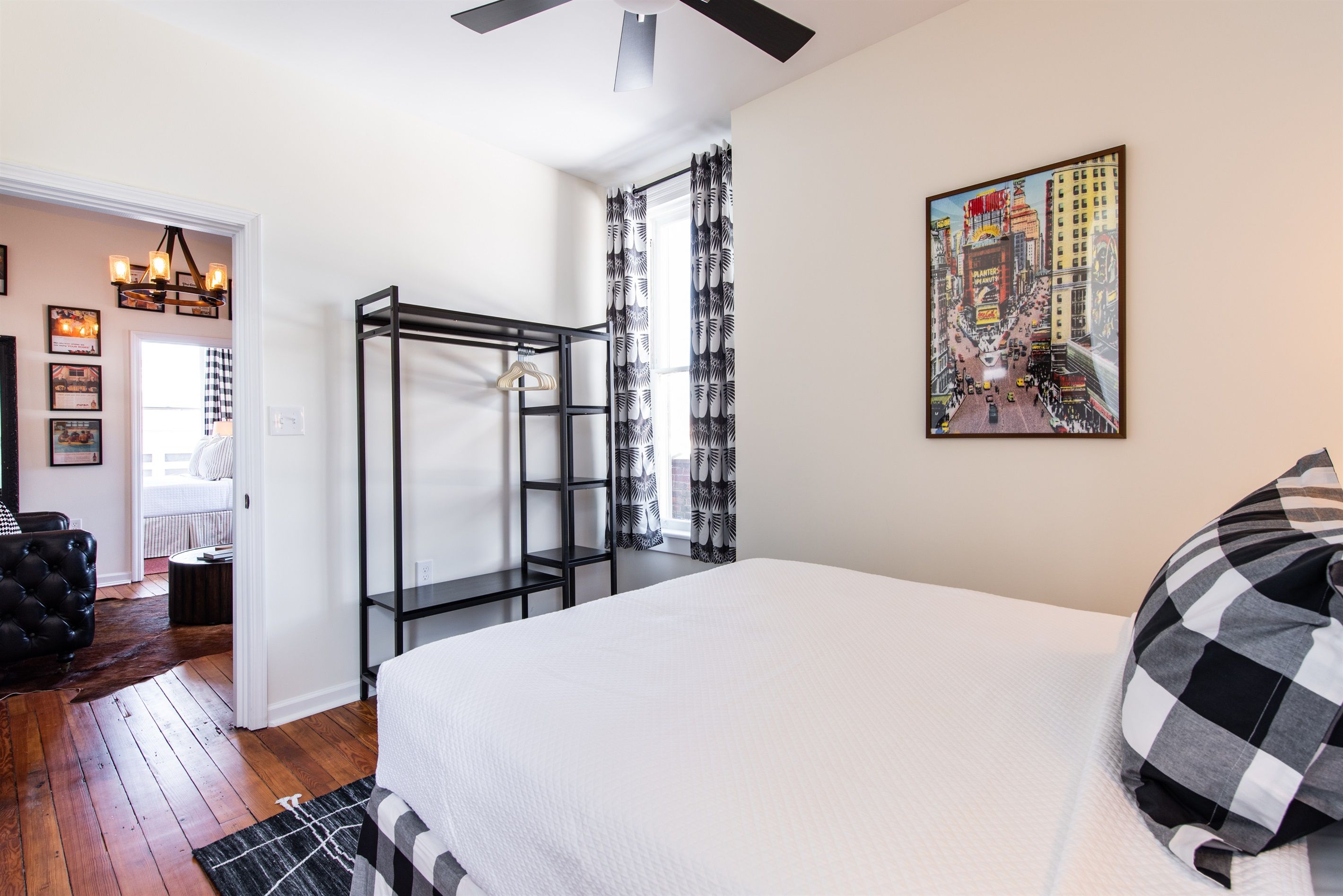 A bedroom with a bed, a black and white checkered pillow, a painting of a cityscape, and a black metal shelf.