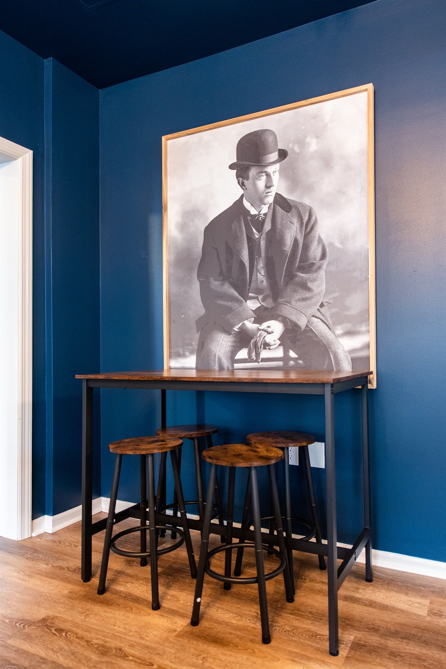 Une pièce avec un mur bleu, une table en bois et trois tabourets, ainsi qu’une photographie en noir et blanc encadrée accrochée au mur.