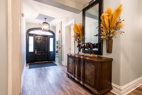 Un pasillo con piso de madera, una puerta oscura, un espejo y un mueble decorativo con un jarrón de plantas secas.