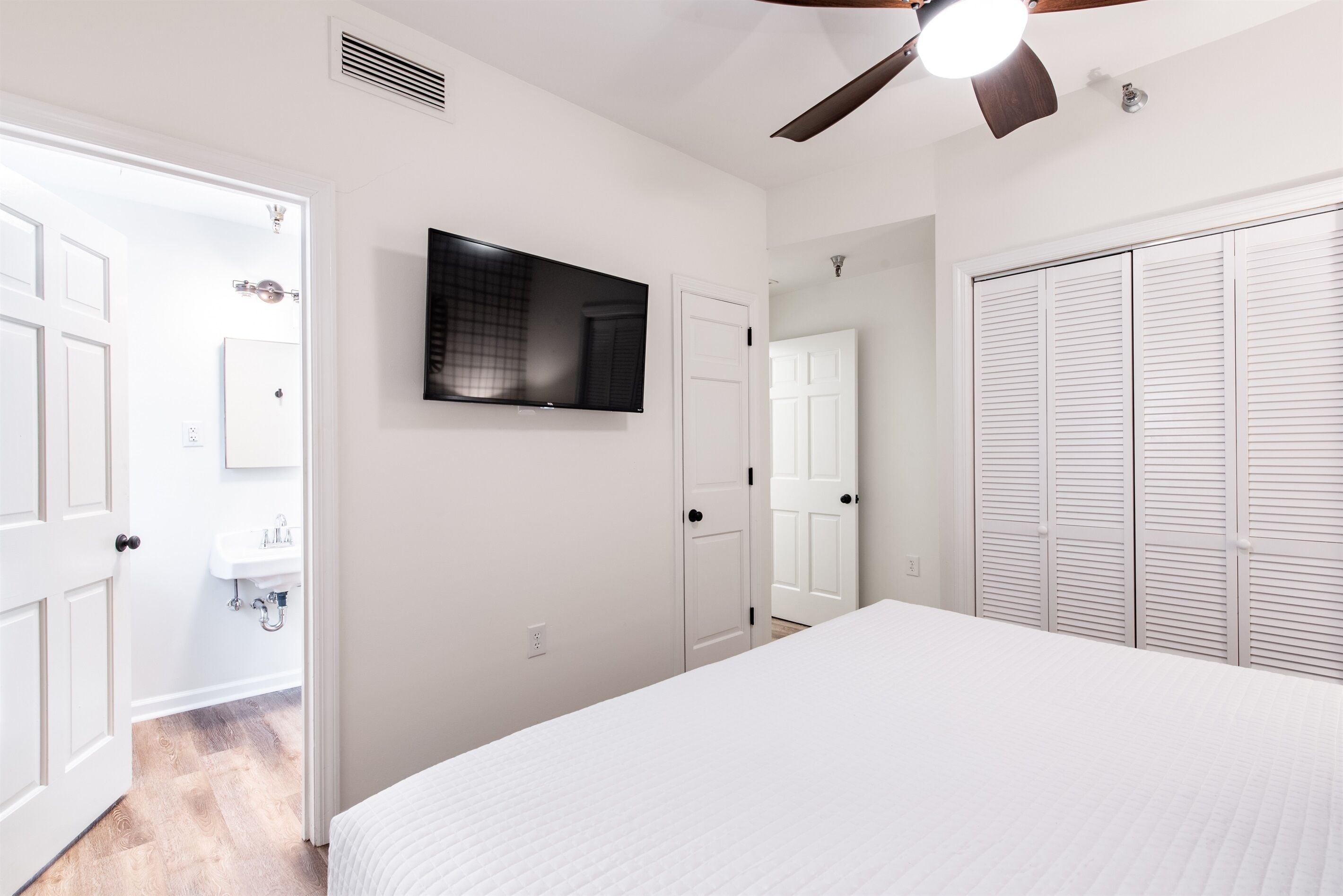 Un dormitorio con cama, ventilador de techo, televisor, armario y baño con lavamanos.