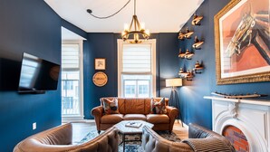 A living room with a brown leather sofa, a coffee table, and a fireplace. There is a television mounted on the wall and a chandelier hanging from the ceiling.