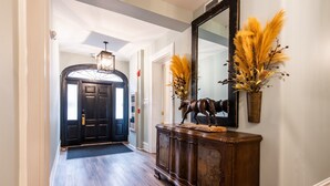 Un pasillo con piso de madera, una puerta oscura, un espejo y un mueble decorativo con un jarrón de plantas secas.