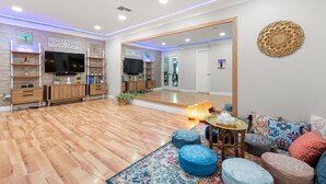 Un salon moderne avec un plancher en bois, un téléviseur, un grand miroir et un coin salon agrémenté de coussins colorés.